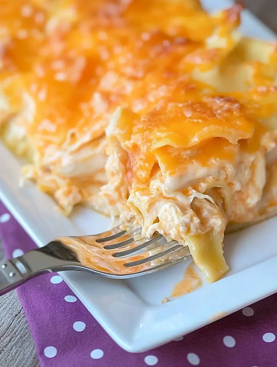 Une fourchette soulève une portion de lasagne au poulet crémeuse et fromagée d'une assiette blanche, avec une serviette à pois violets dessous.