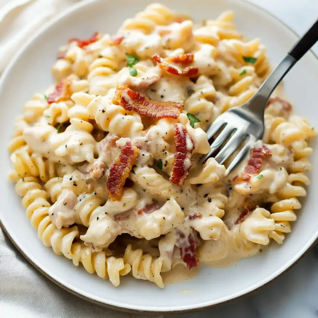 Une assiette de pâtes crémeuses avec des morceaux de bacon et une garniture verte, servie avec une fourchette.