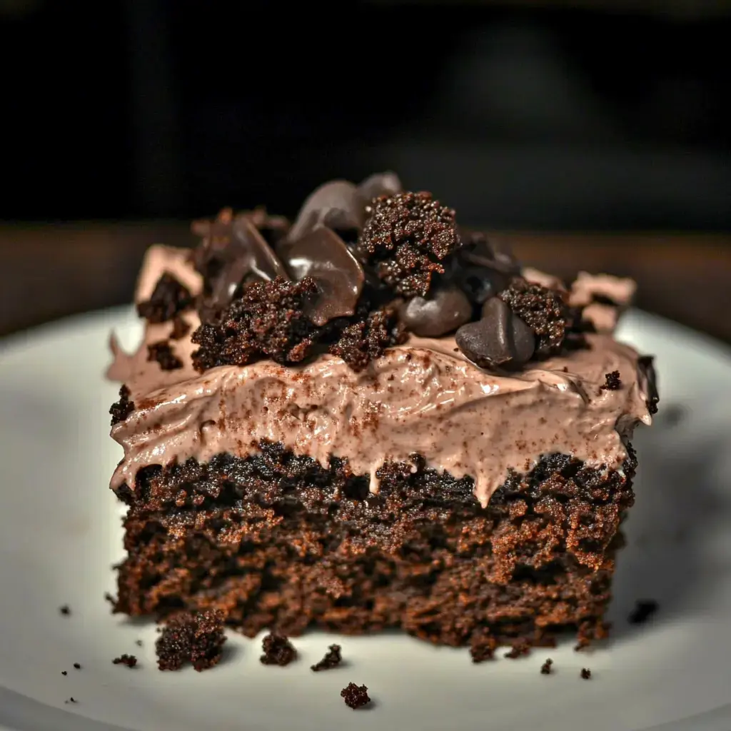 Une part de gâteau au chocolat nappée de fudge et parsemée d’éclats de chocolat sur une assiette blanche.