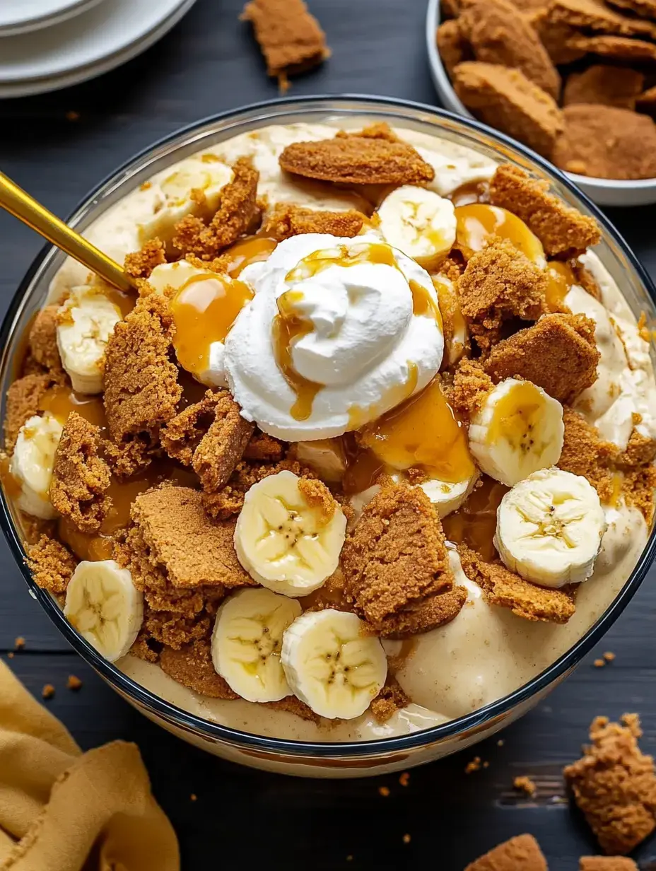 Un bol en verre rempli de dessert en couches garni de tranches de banane, biscuits émiettés, crème fouettée et sauce caramel.