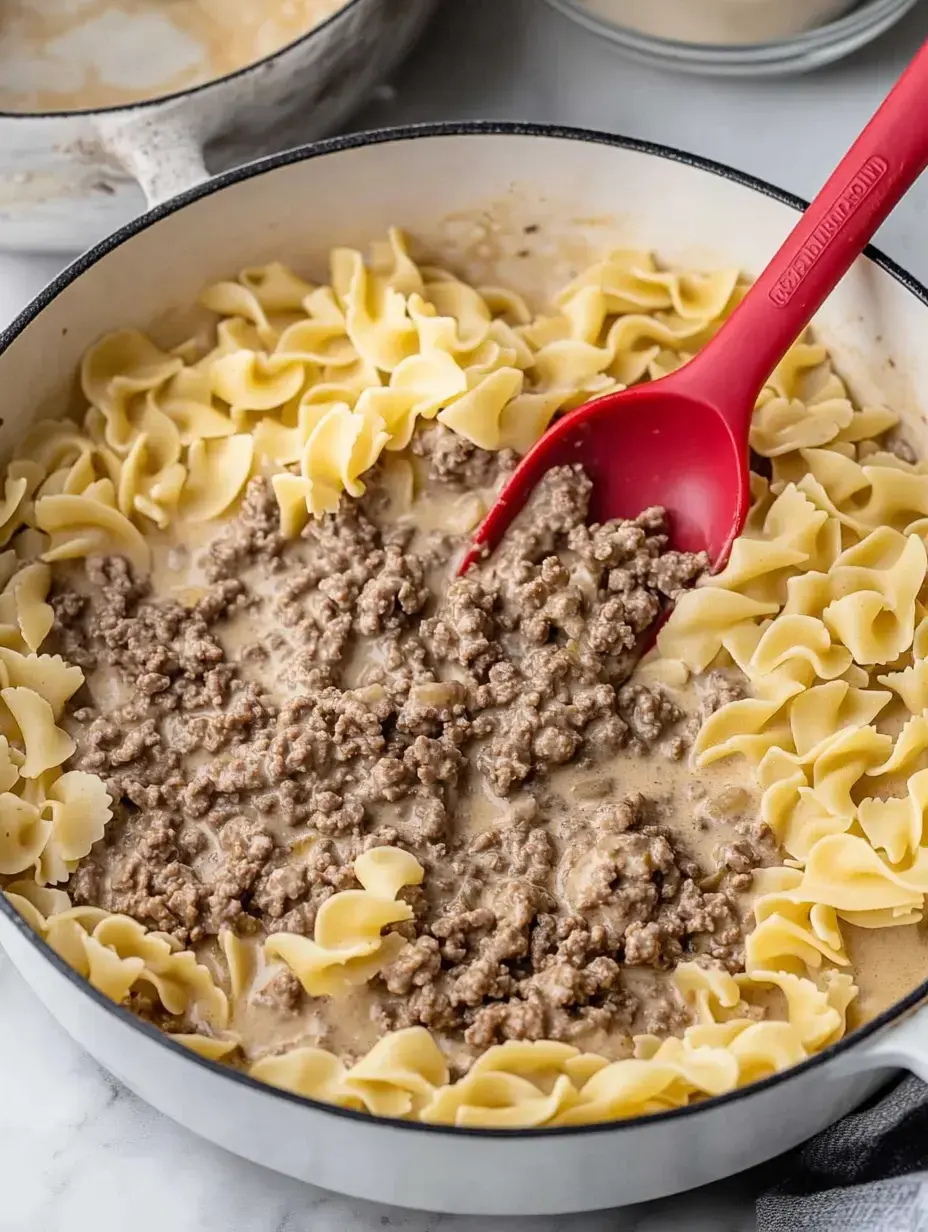 Une poêle remplie de nouilles aux œufs cuites et de bœuf haché dans une sauce crémeuse, avec une spatule rouge mélangeant les ingrédients.