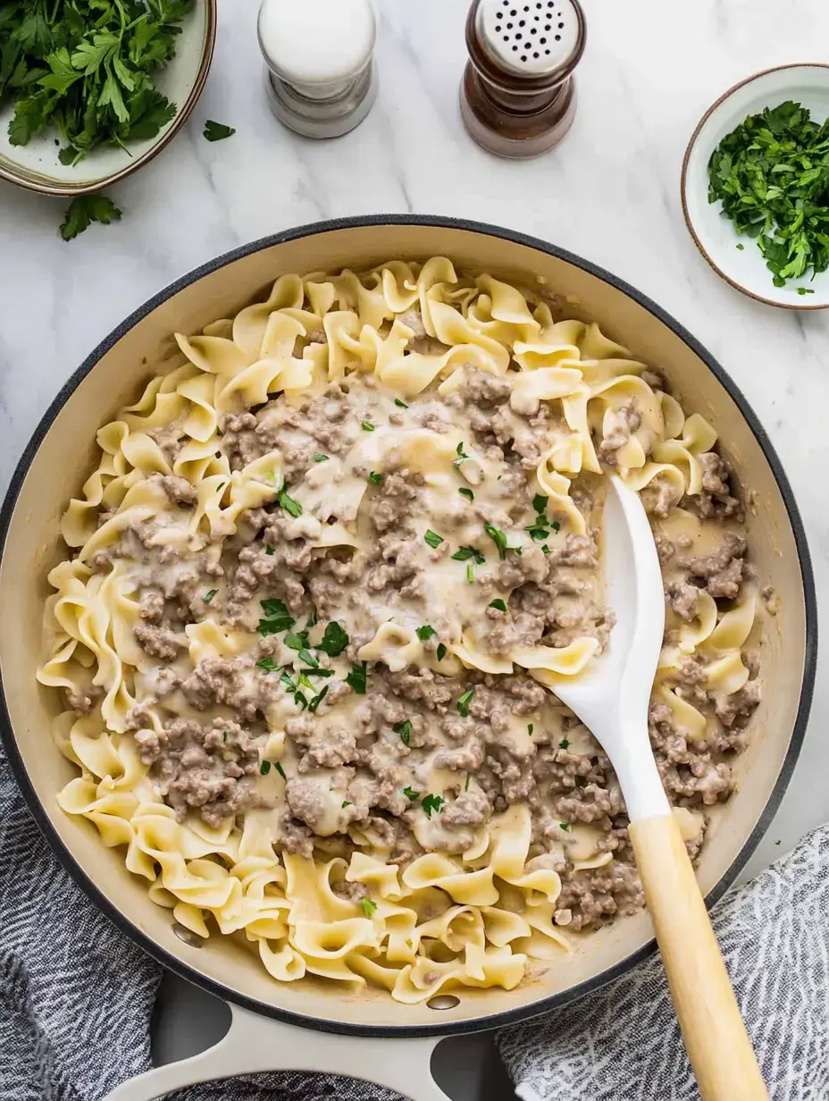 Un plat crémeux de bœuf et nouilles garni de persil, servi dans une grande poêle avec des herbes fraîches et des condiments.